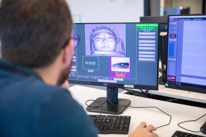 The back of Freek van Ede's head is in the foreground of the picture, blurry and out of focus. The focus of the picture is on the computer screen that he is staring at, which displays a black and white image of the electroencephalogram (EEG) experiment participant's face. There is a red circle around the participant's left eye, and below the image of the face, there is a zoomed in image of the eye.